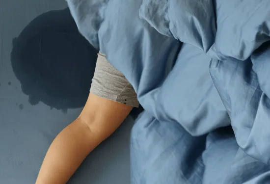 Person in bed, leg extending out from beneath blue comforter with a big wet spot on the sheets